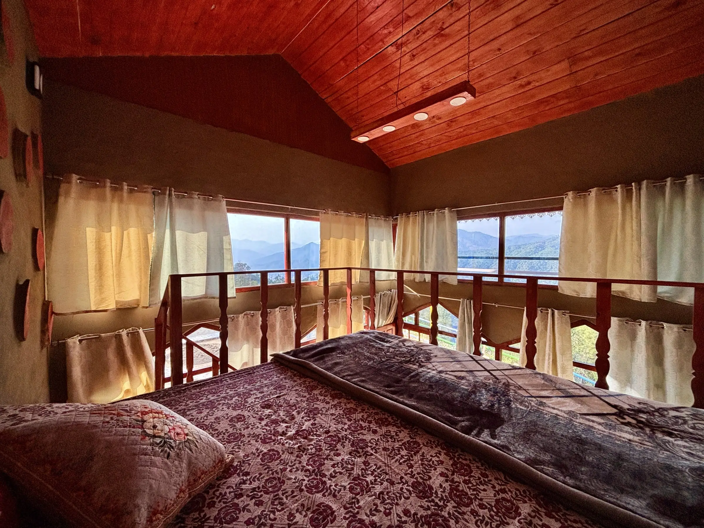 A railed sleeping platform under a wooden ceiling, windows onto hills
