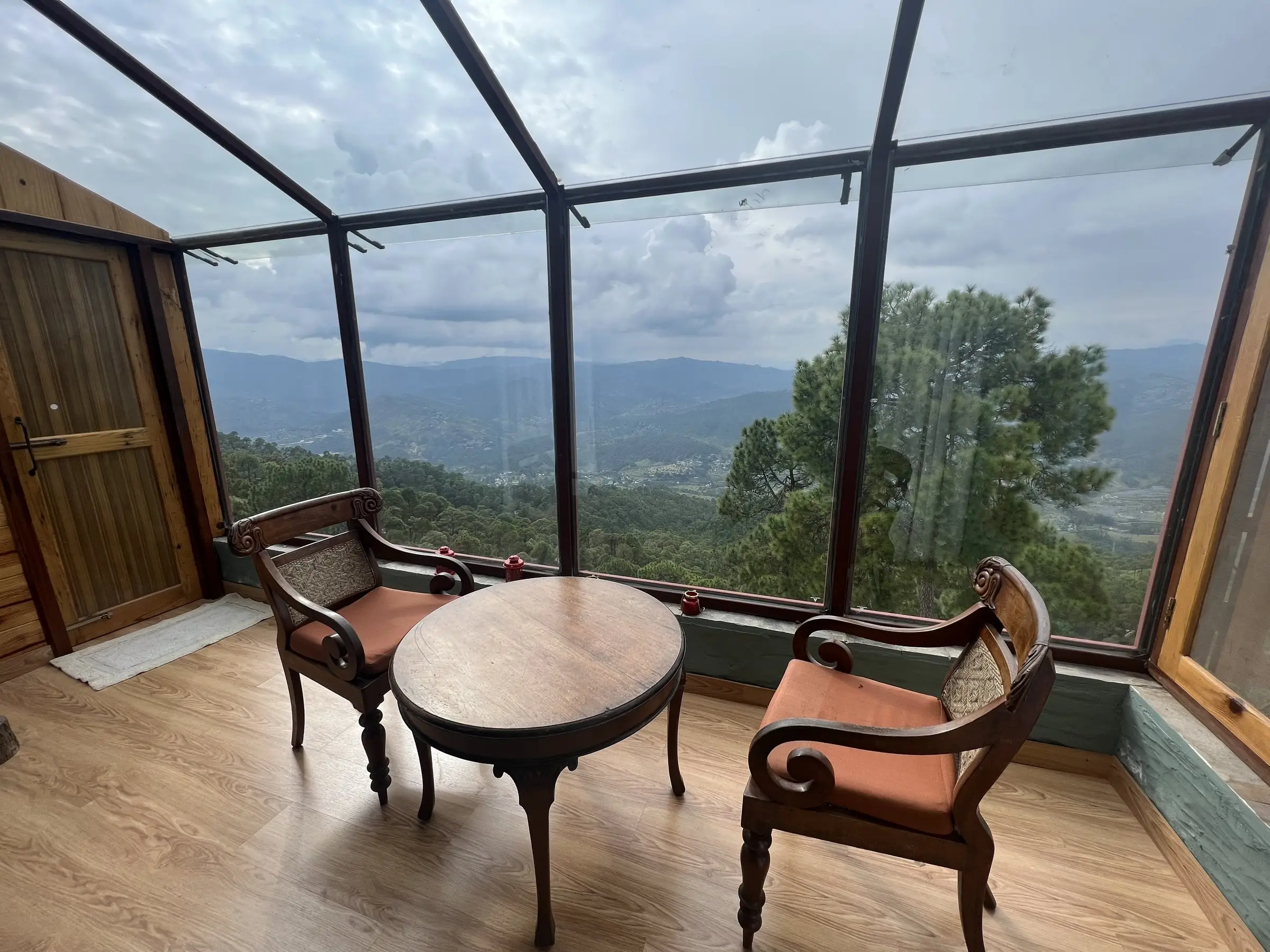 A glass-walled corner with a round table and two chairs over a forested valley