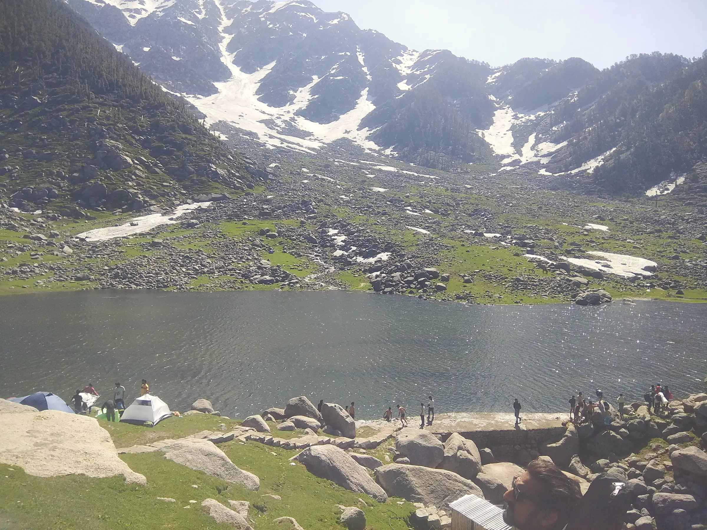 A mountain lake with snow on the slopes above and tents on the shore