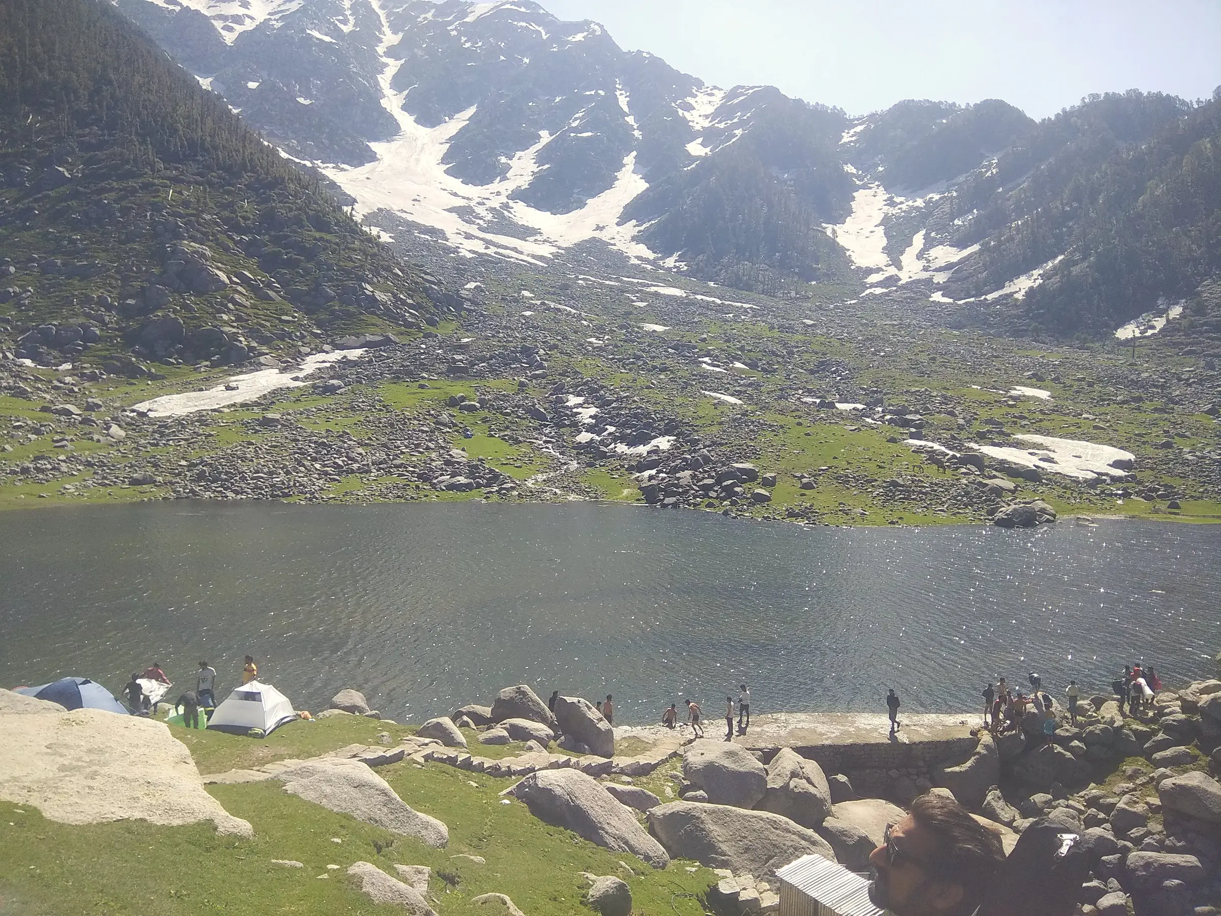 A mountain lake with snow on the slopes above and tents on the shore