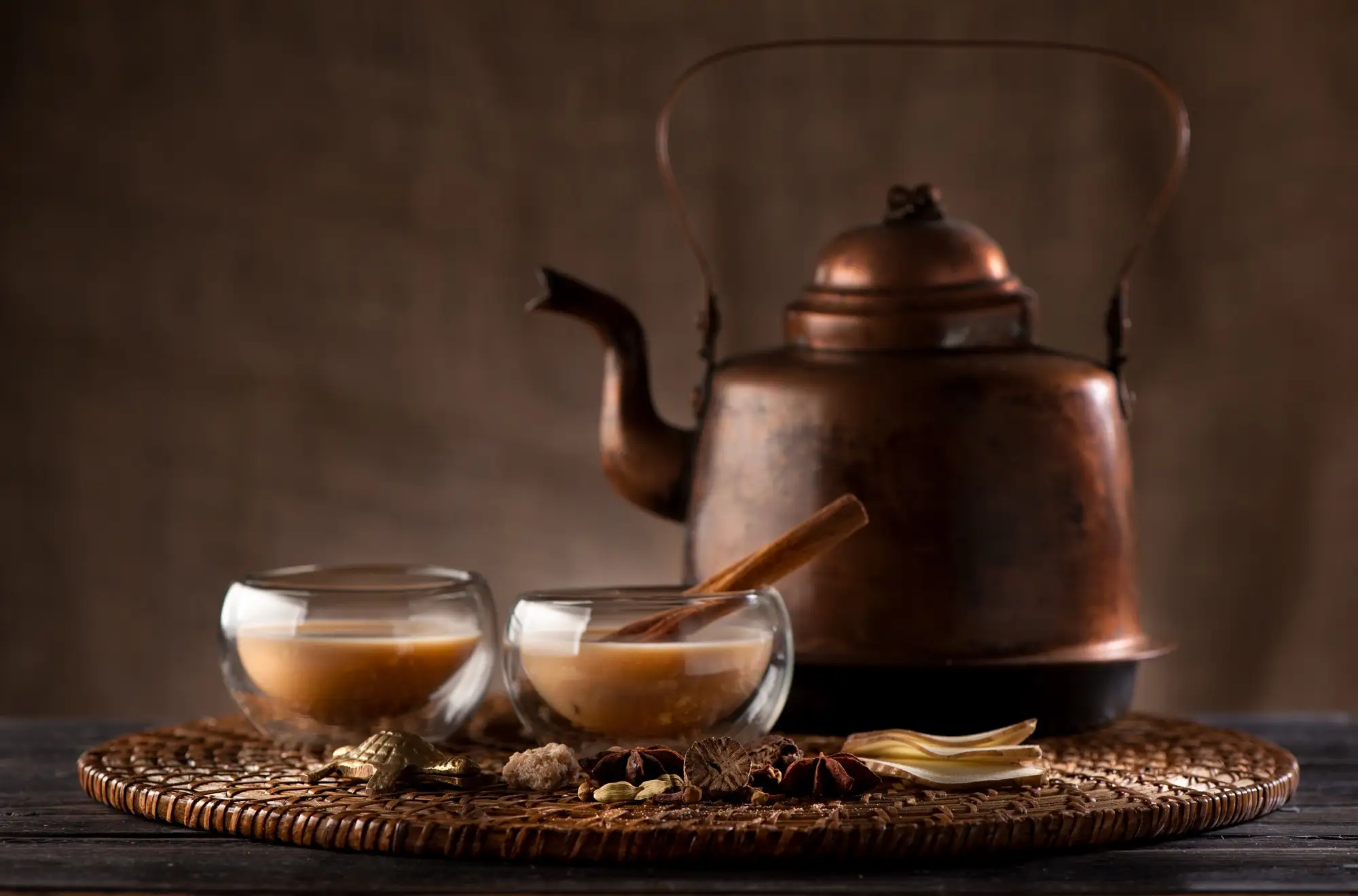 A copper kettle and two glasses of chai on a woven tray