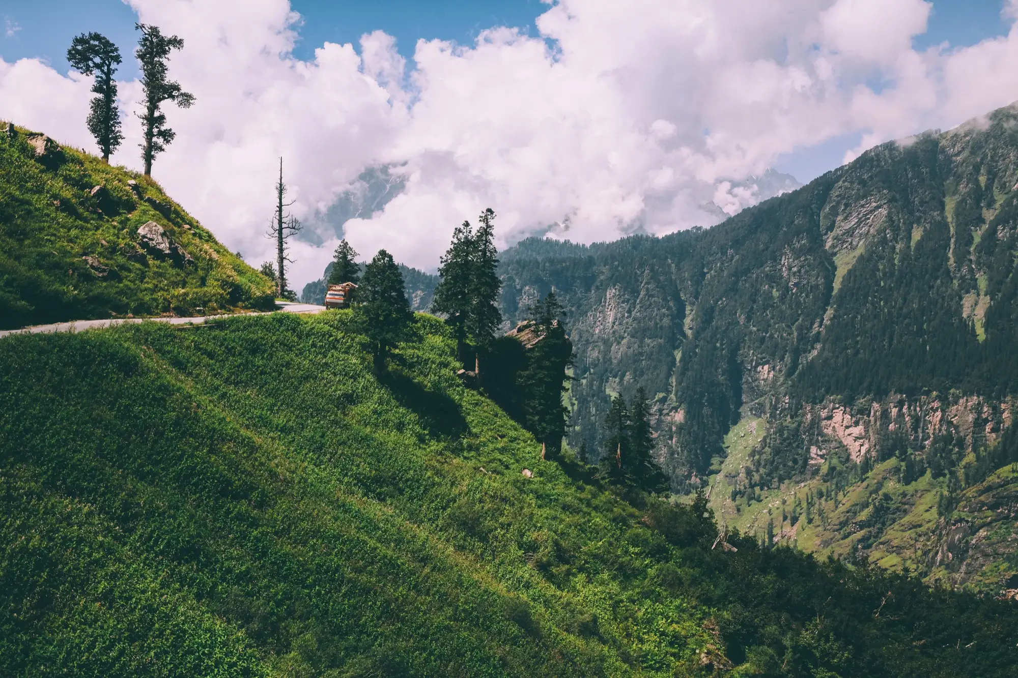 A road curving along a steep green slope above a deep valley, with a truck on the bend