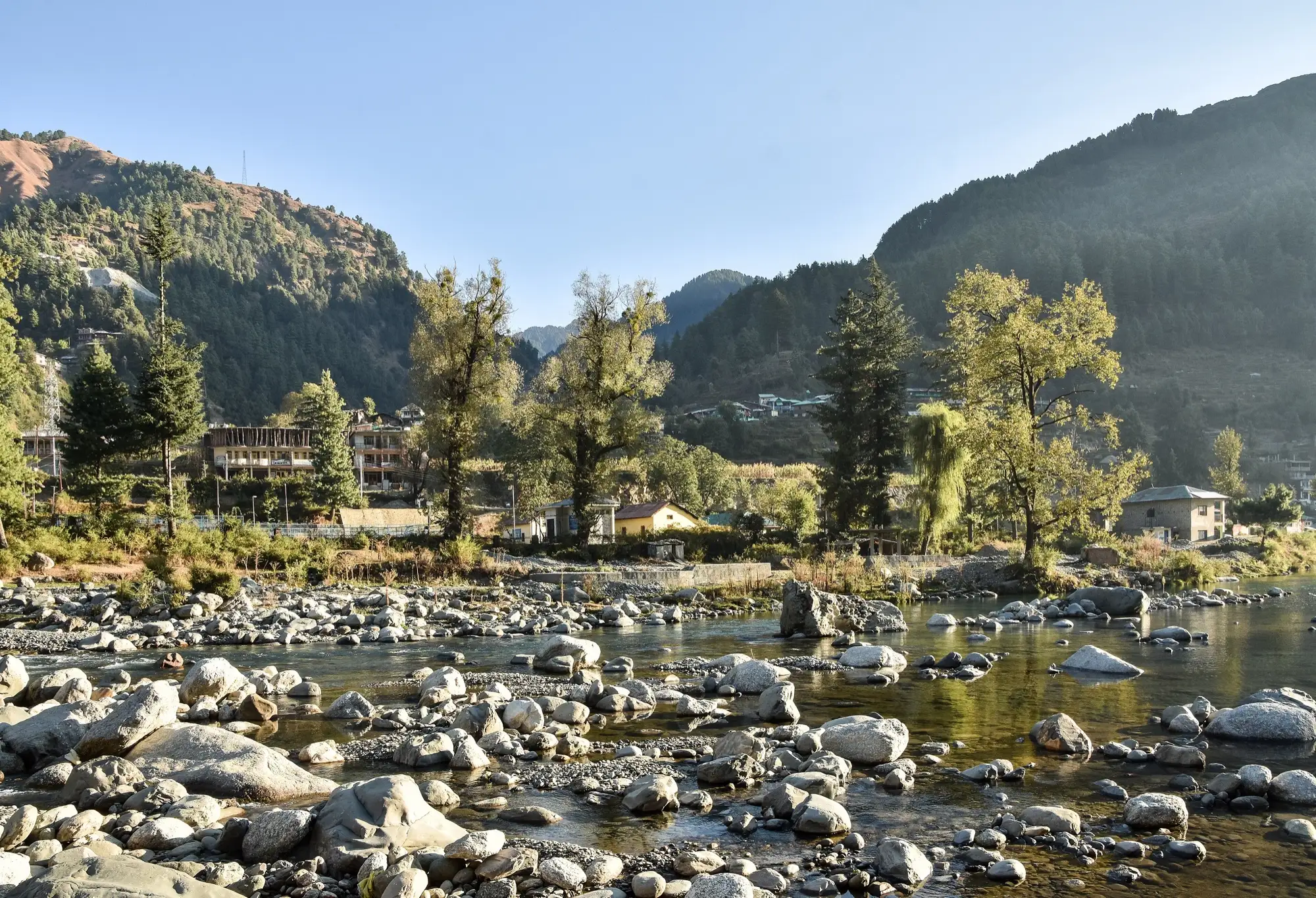 The Uhl river over rounded stones at Barot, with forested slopes and village houses behind